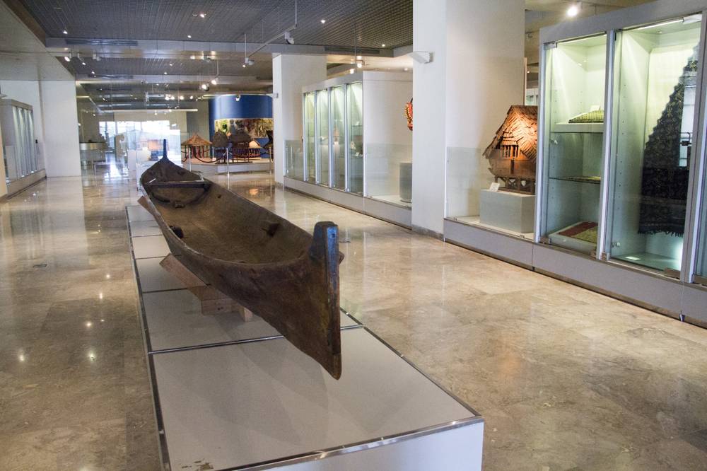 A traditional wooden outrigger boat is displayed in a bright, modern museum gallery.