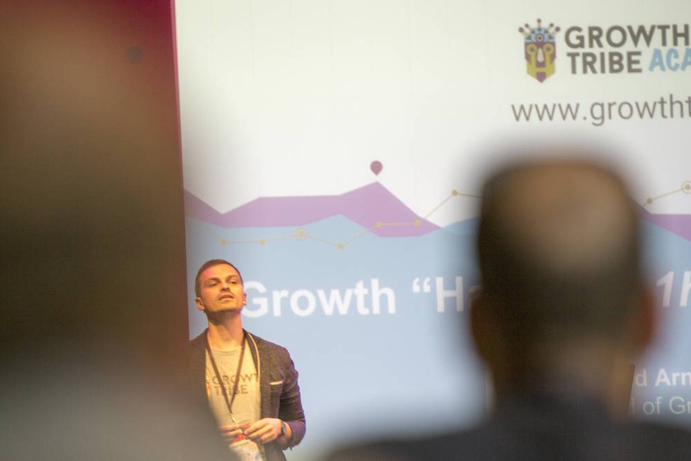 A speaker stands beneath a screen reading 'Growth Hacking 1hr Power Session' as an audience member watches from behind.