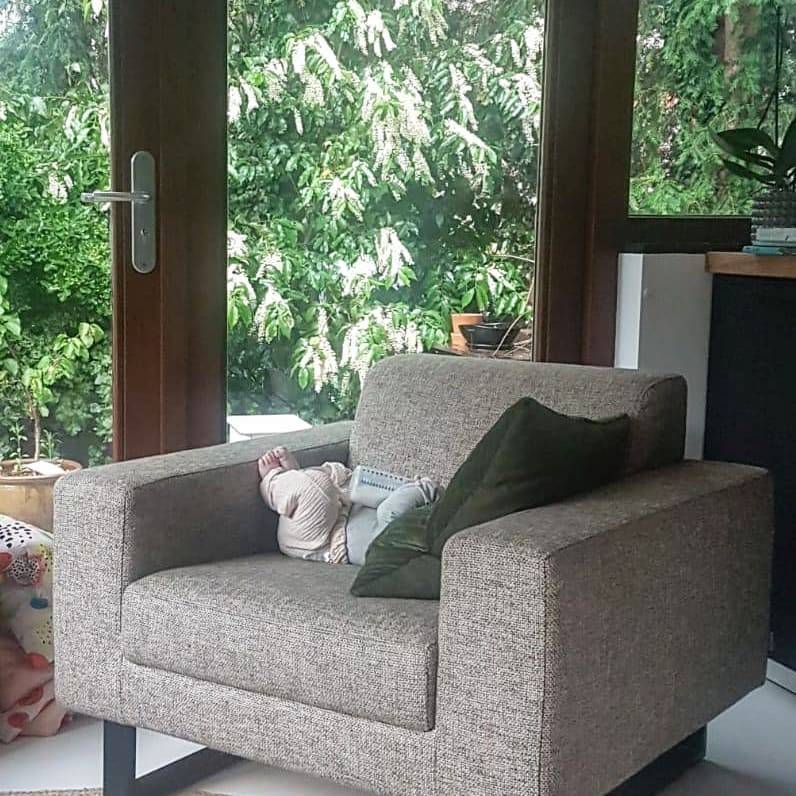 A grey armchair sits by large windows with a baby lying on it, greenery visible outside.