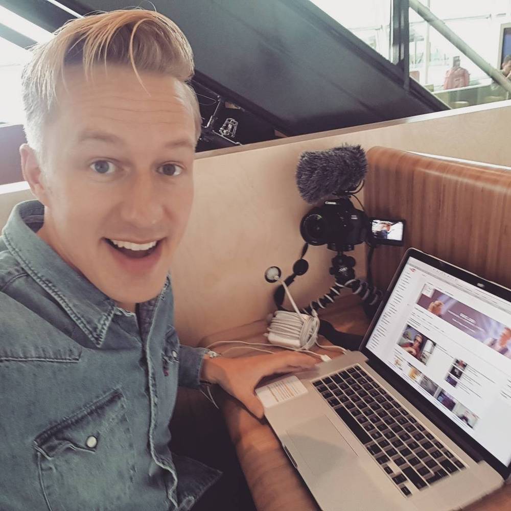 A man films himself with a smartphone rig while working on a laptop at a café table.