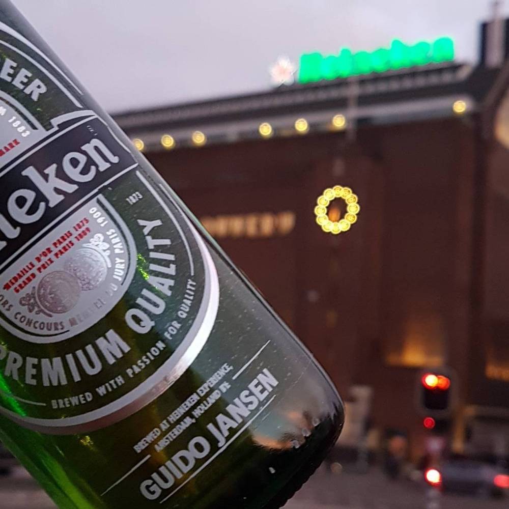 A Heineken beer bottle personalized with the name Guido Jansen, held against the lit Heineken brewery building at dusk.