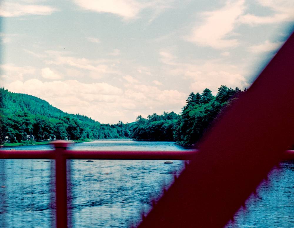 A wide river flows between tree-covered banks, viewed from a bridge with a large red structural beam in the foreground.
