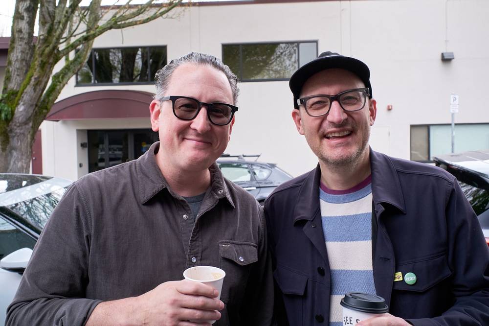 Two men smile for the camera outdoors, each holding a coffee cup. Both wear glasses; one is in a corduroy shirt, the other in a dark jacket with a striped sweater and a black cap.