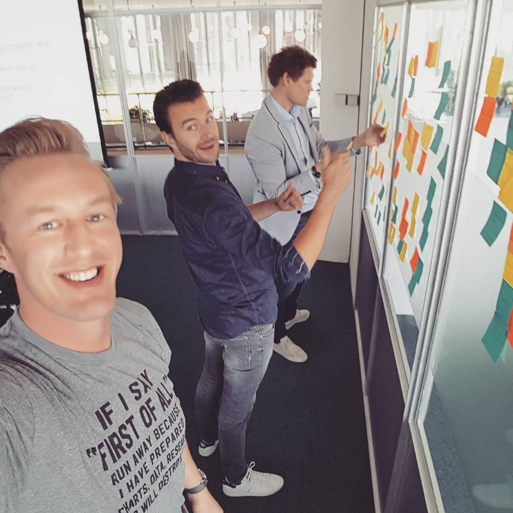 Three colleagues smiling next to a wall covered in colorful sticky notes during a brainstorm session.