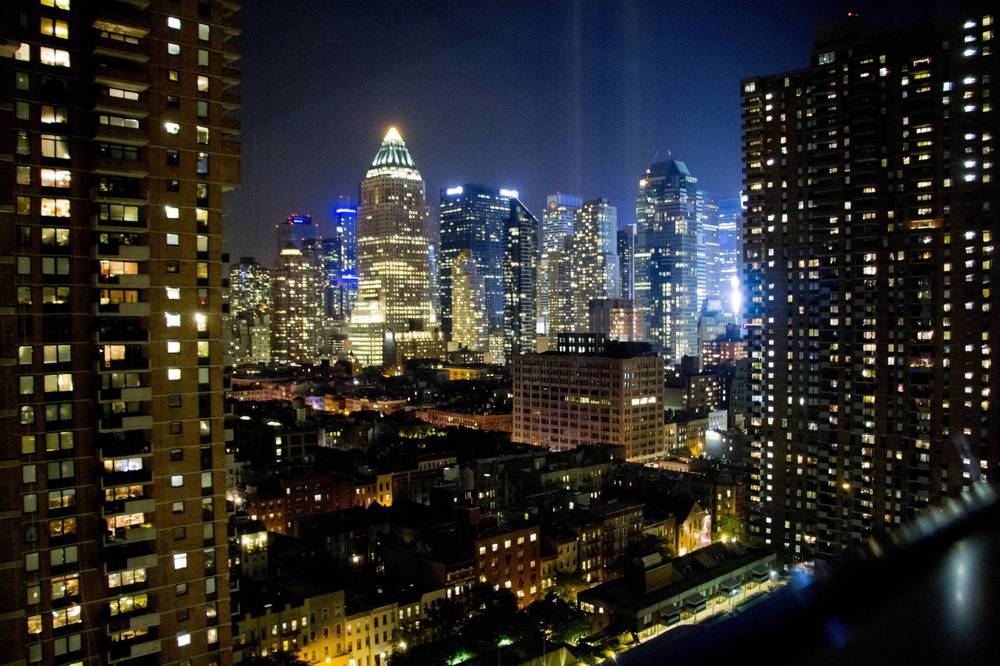 City lights fill the Manhattan skyline at night, seen from a high apartment window.