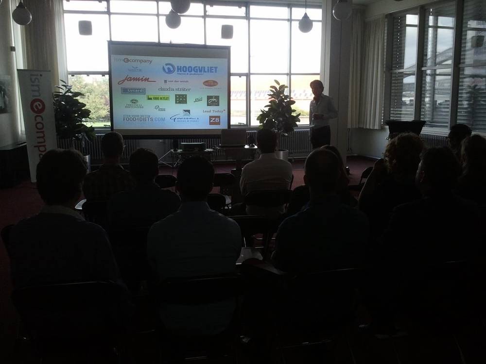 An audience watches a presenter beside a slide of retail brand logos.