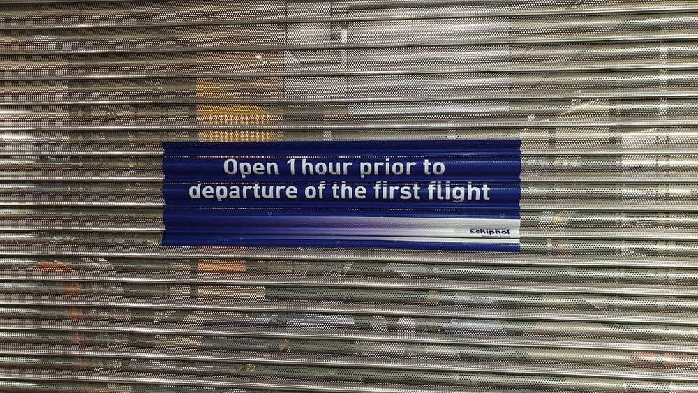 A closed metal shutter with a sign reading "Open 1 hour prior to departure of the first flight" at Schiphol.