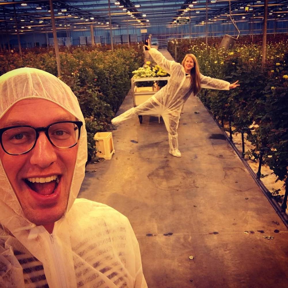 A man in glasses and a woman, both wearing white disposable coveralls, posing playfully in a greenhouse aisle.