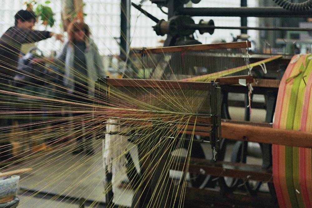 A weaving loom in a sunlit studio, with hundreds of warp threads in yellow, green, and pink fanning out from a striped fabric beam toward the camera; two blurred figures stand in conversation in the background.