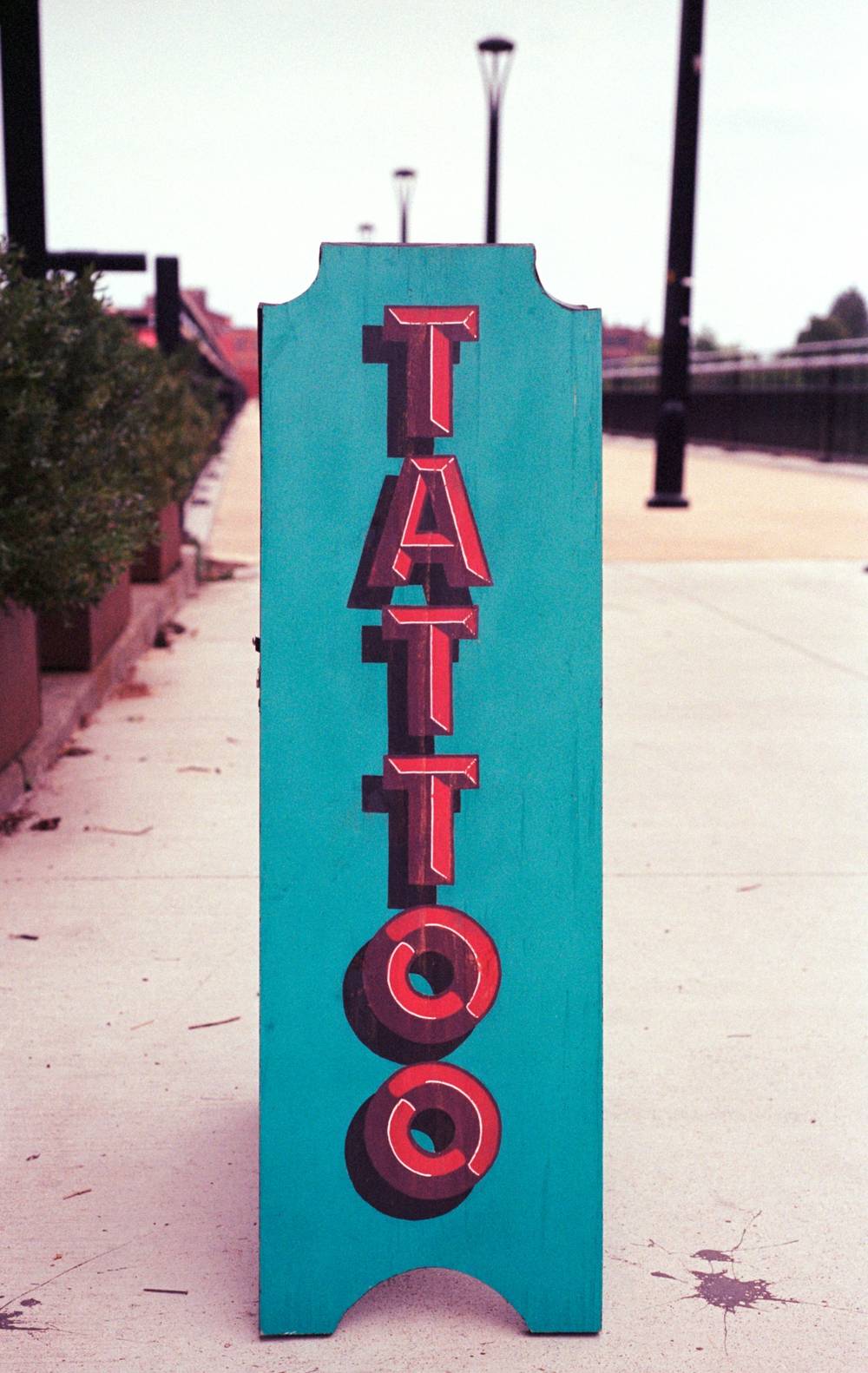A turquoise vertical sandwich board on a concrete sidewalk reads TATTOO in red block letters with black drop shadows.