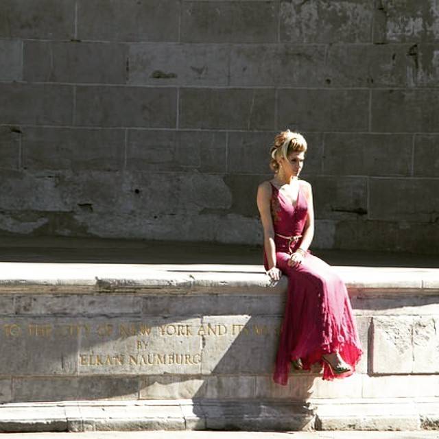 A woman in a red dress sits on a stone ledge in front of an engraved wall.
