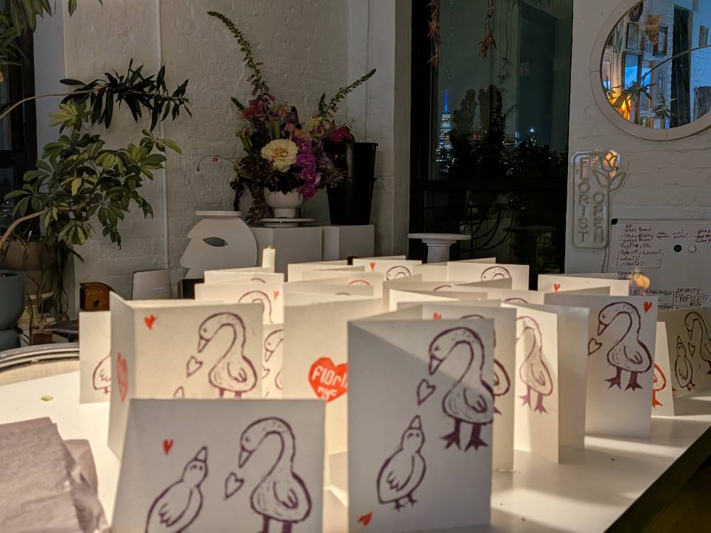 Zoomed out version of mother's day prep display. More of the cards are seen. Mother goose and baby goose on cards with hearts. An arraignment of flowers and windownin background