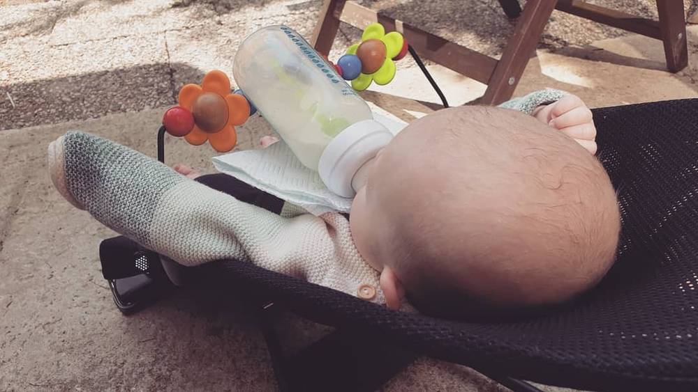 A baby lies in an outdoor bouncer chair with toy attachments and a bottle nearby.
