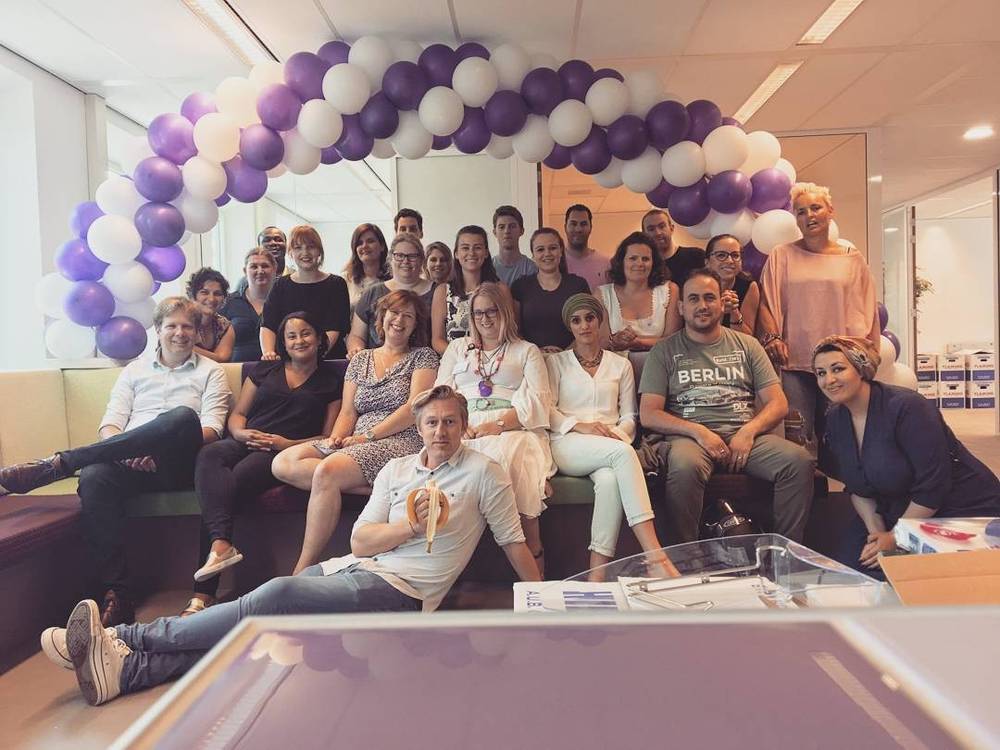 A large group of colleagues posing together under a purple and white balloon arch in an office.