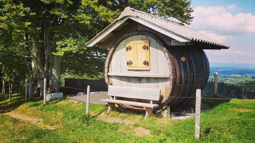 A small wooden barrel-shaped hut with shutters and a bench, set in a grassy alpine meadow.