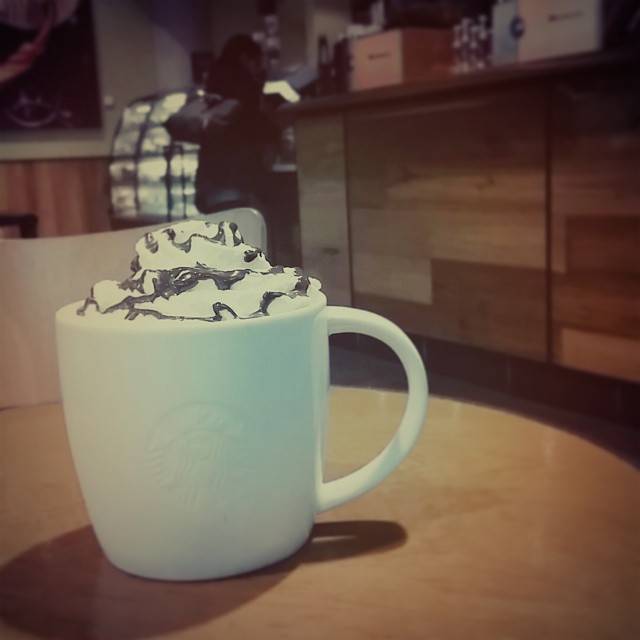 A Starbucks mug topped with whipped cream and chocolate drizzle on a wooden table, with a person visible in the background.