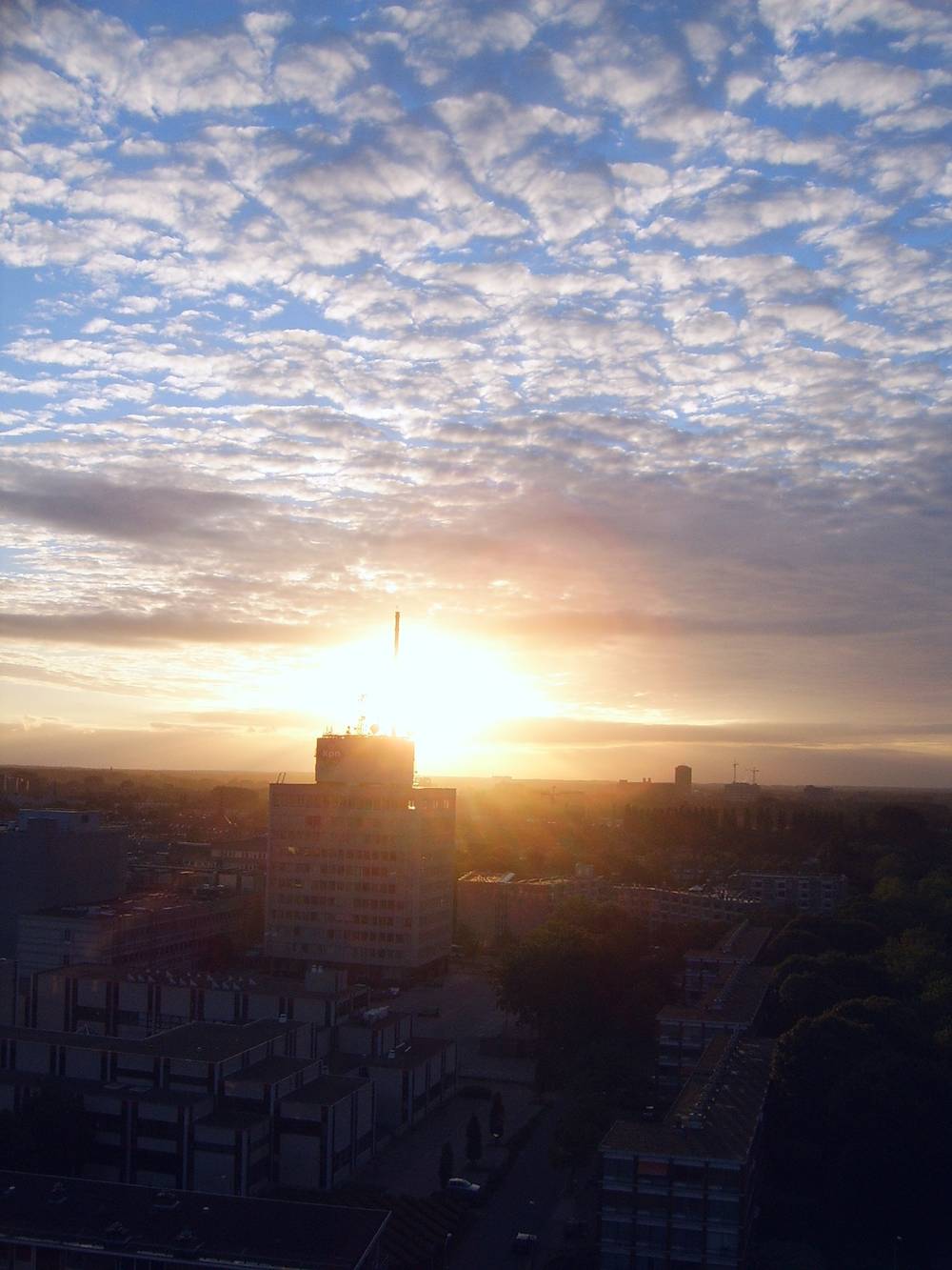 The sun sets in a haze behind a tall tower on the Utrecht skyline, clouds tinted pink.