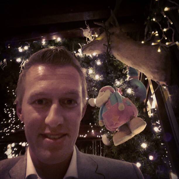 A man's face lit by string lights next to hanging Christmas ornaments and a small plush toy.