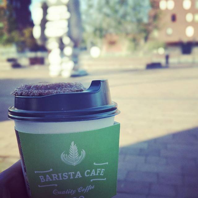 A takeaway coffee cup from Barista Cafe held outdoors on a paved square.