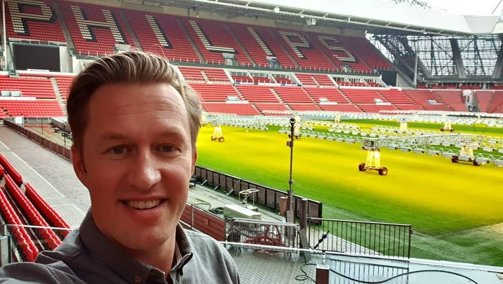 A man takes a selfie in an empty football stadium with red seats, groundskeeping equipment on the pitch.