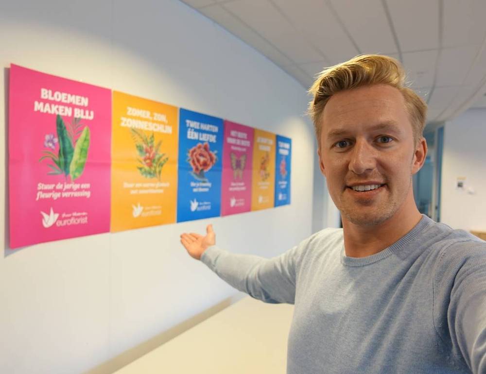 A man gestures toward a row of colorful flower-themed posters on an office wall.