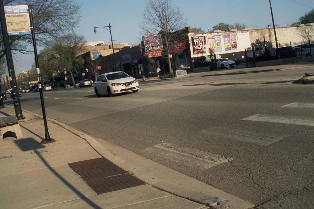 A white Nissan Sentra speeds into a crosswalk past a "74 Fullerton" CTA bus stop sign, with a hand car wash and window tint shops in the background.