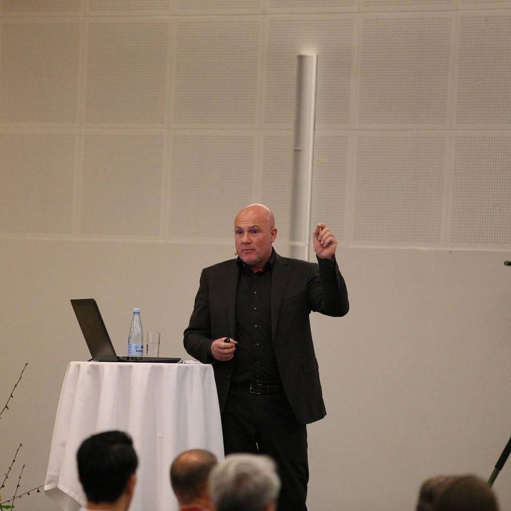 A man in a black suit gestures while presenting from a small podium on stage.