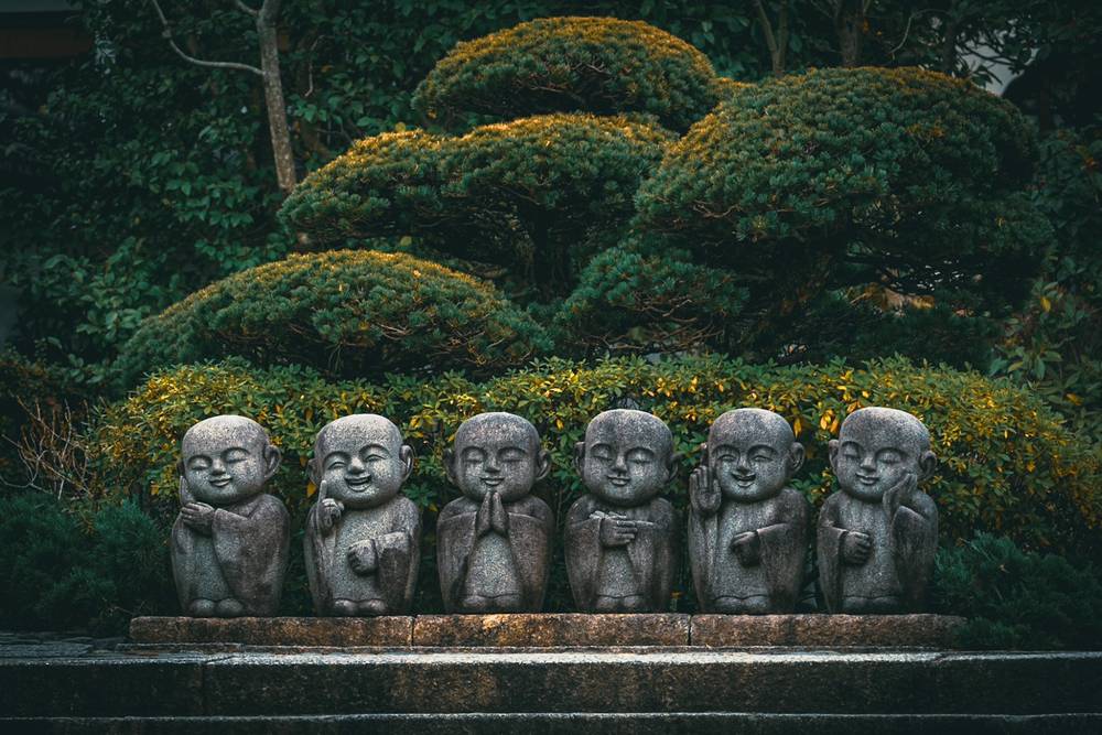 A row of 6 child-like Buddha statues carved from stone along the top of a short wall in front of a manicured garden at the entrance of Reigen-in Temple, near Tofuku-ji in Kyoto Japan.