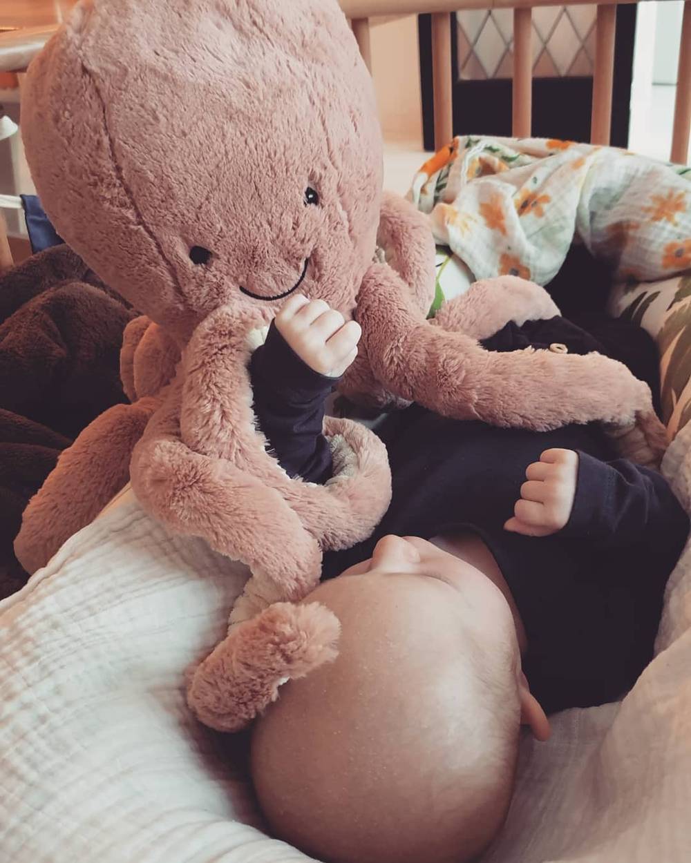 A baby holds a plush pink octopus toy while lying on a blanket.