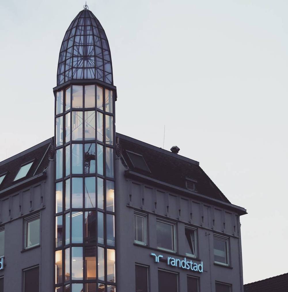 A narrow historic building topped with a glass-enclosed spire, with a Randstad sign at street level.