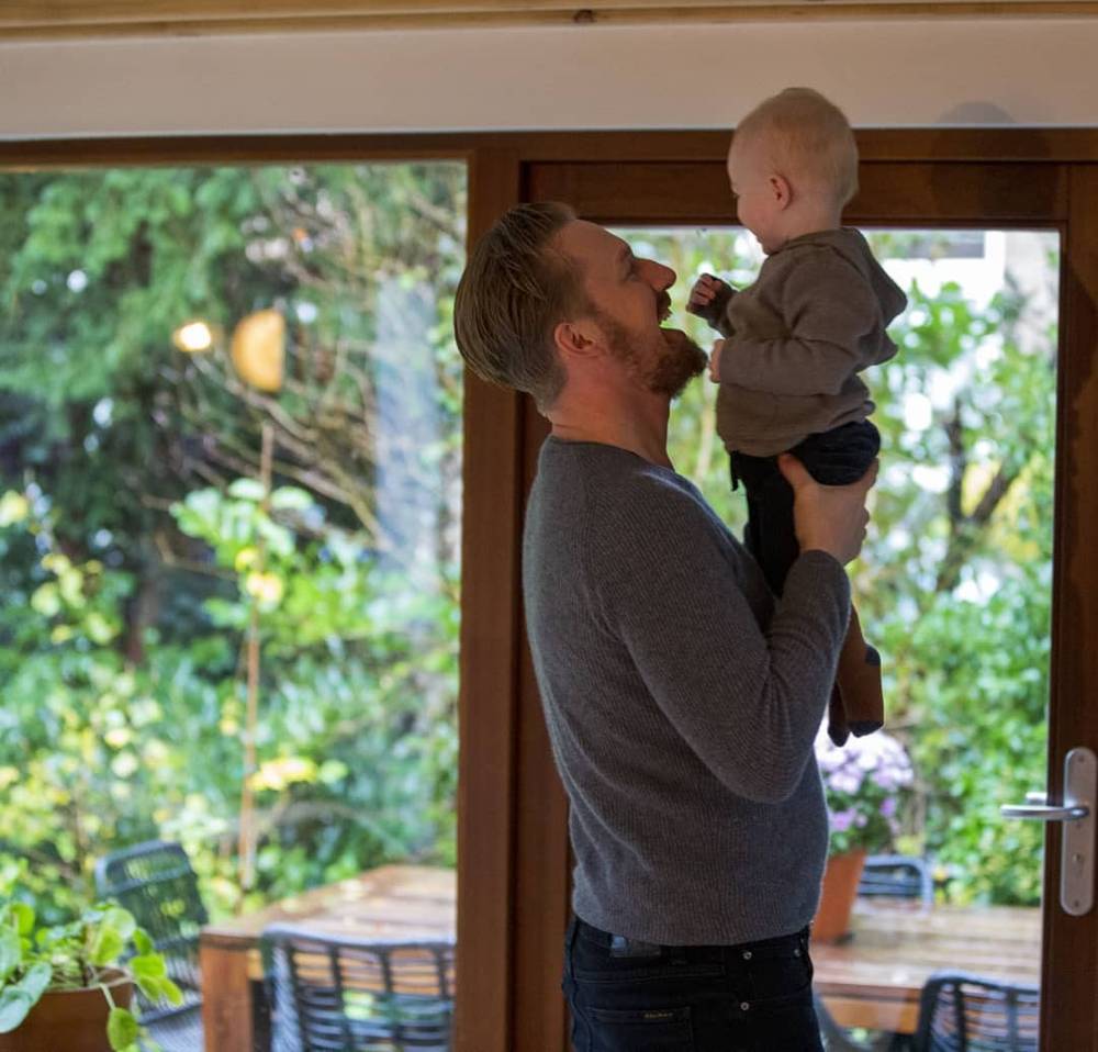 A man lifts a toddler up to kiss him near an open glass door with greenery outside.