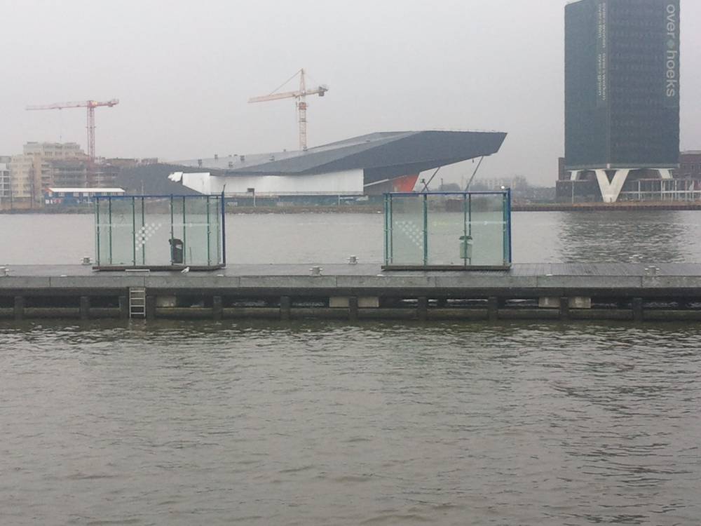 A distinctive angular building juts out over the water next to construction cranes on an overcast day.