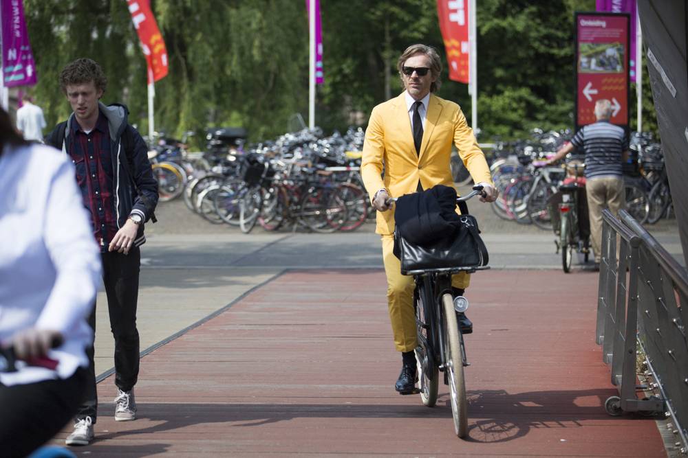 A man in a mustard yellow suit cycles along a path lined with parked bicycles near the venue.