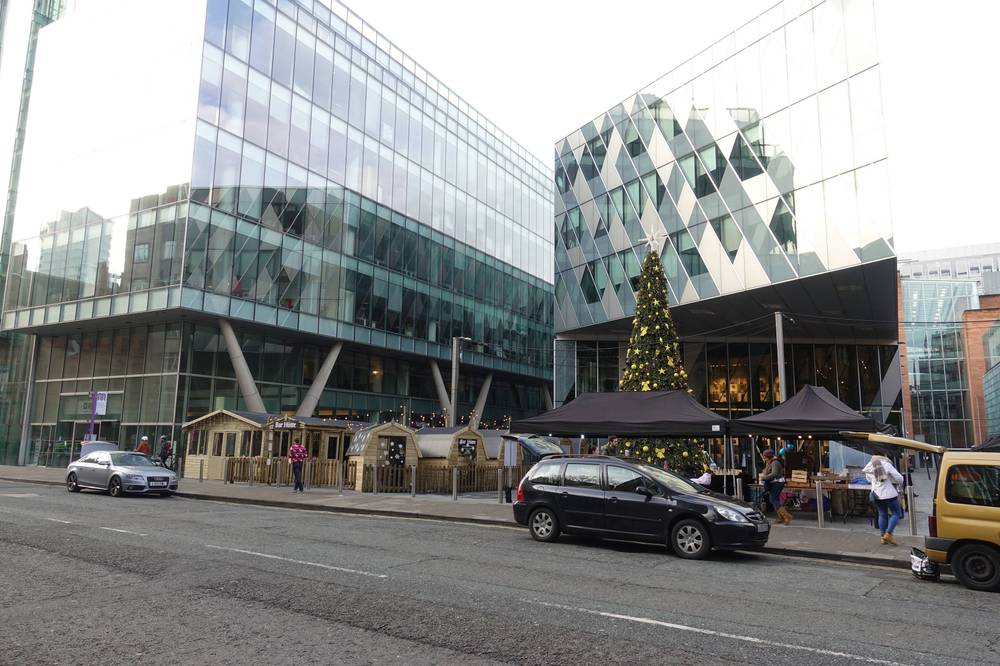 Modern glass office buildings and a lit Christmas tree stand on a Manchester street.