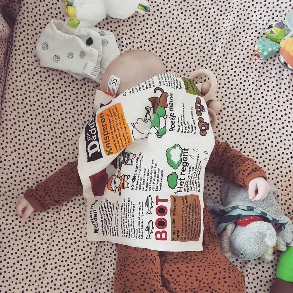 A newborn lies on a patterned blanket wearing a paper hat made from a Dutch newspaper, surrounded by toys.
