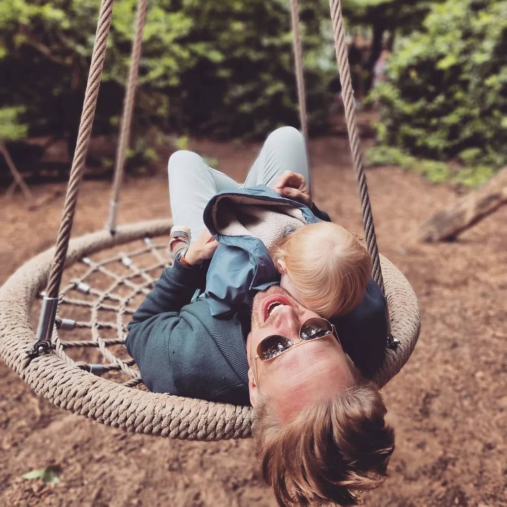 A man lying on a round hanging swing chair, playfully nose-to-nose with a toddler leaning over him.