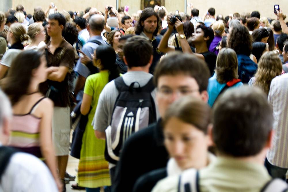 A dense crowd of tourists, many holding cameras, packs a gallery at the Louvre.