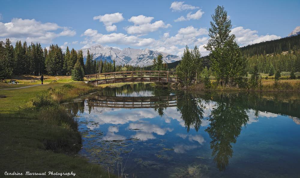 A view of Cascade Ponds, in Alberta, Canada. In the foreground, water with nice reflections. Behind, a bridge with some trees. In the distance, mountains.
