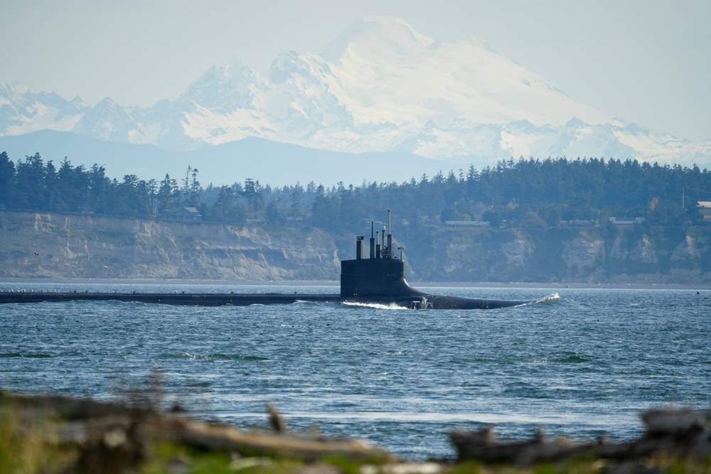Subject is a submarine on the surface with a snow-covered peak, Mt Baker, in the background.  The foreground is grass and driftwood.