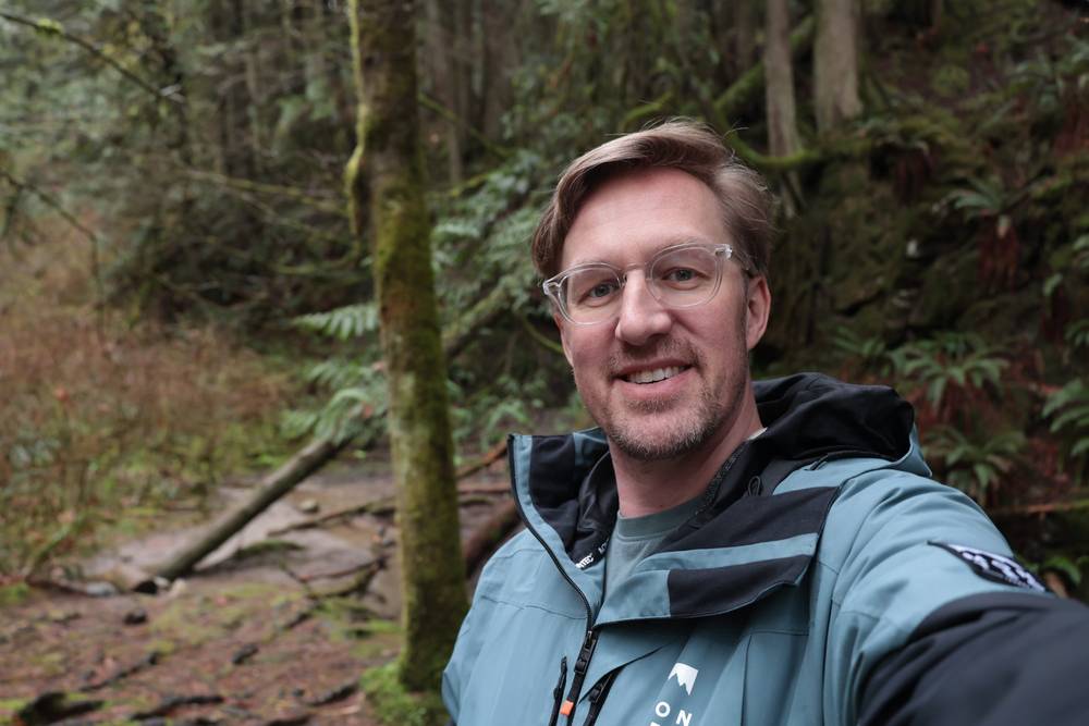A man wearing glasses and a jacket takes a selfie in a mossy forest.