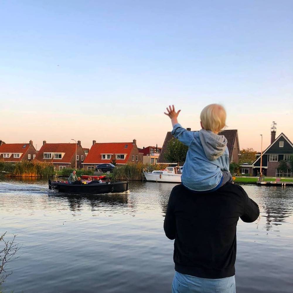 A toddler perched on an adult's shoulders waves toward a small Dutch village with red-roofed houses and a moored boat.