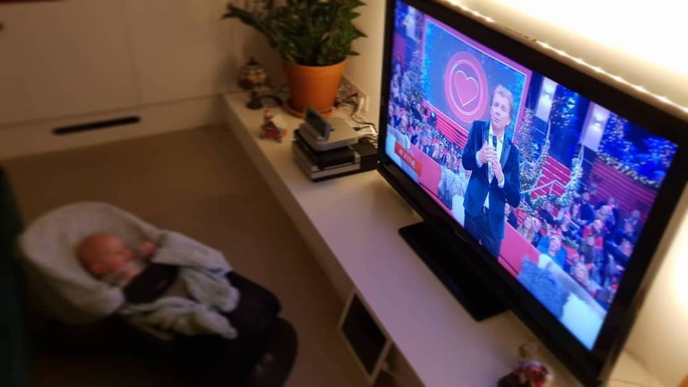 A newborn lies on a low shelf near a television playing a Dutch broadcast.