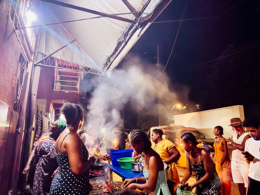 Scène de street food dans les rues de Androany : des femmes malagasy préparent des Masikita, des brochettes de zébu marinées et les font cuire sur un barbecue pendant que les clients attendent