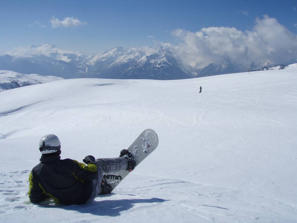 A snowboarder lying back in the snow on an open slope, mountains stretching into the distance.