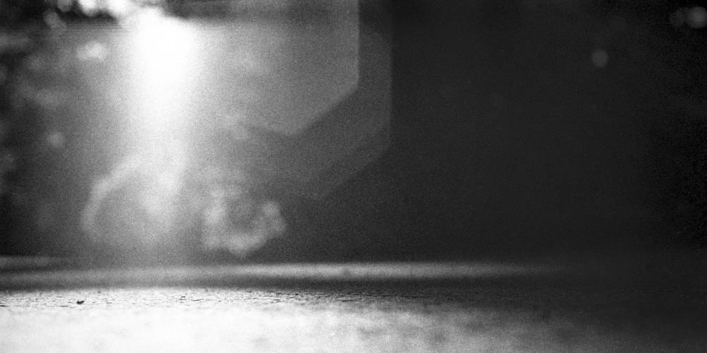 An abstract black and white scene with multiple layers of lens flare in the upper left fading to darkness on the right. The lower left is a well-lit cracked surface with very shallow depth of field revealing it only partially.