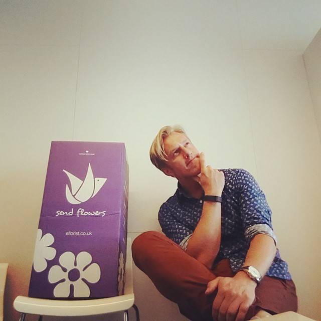 A man crouches thoughtfully beside a purple box branded 'send flowers, eflorist.co.uk'.