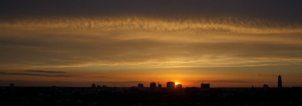 A wide sunset over the Utrecht skyline seen from a rooftop, with rows of houses in the foreground and church towers on the horizon.
