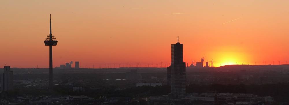 Sunset. The skyline of a major city with its television tower, an office building, and coal-fired power plants and wind turbines in the background.