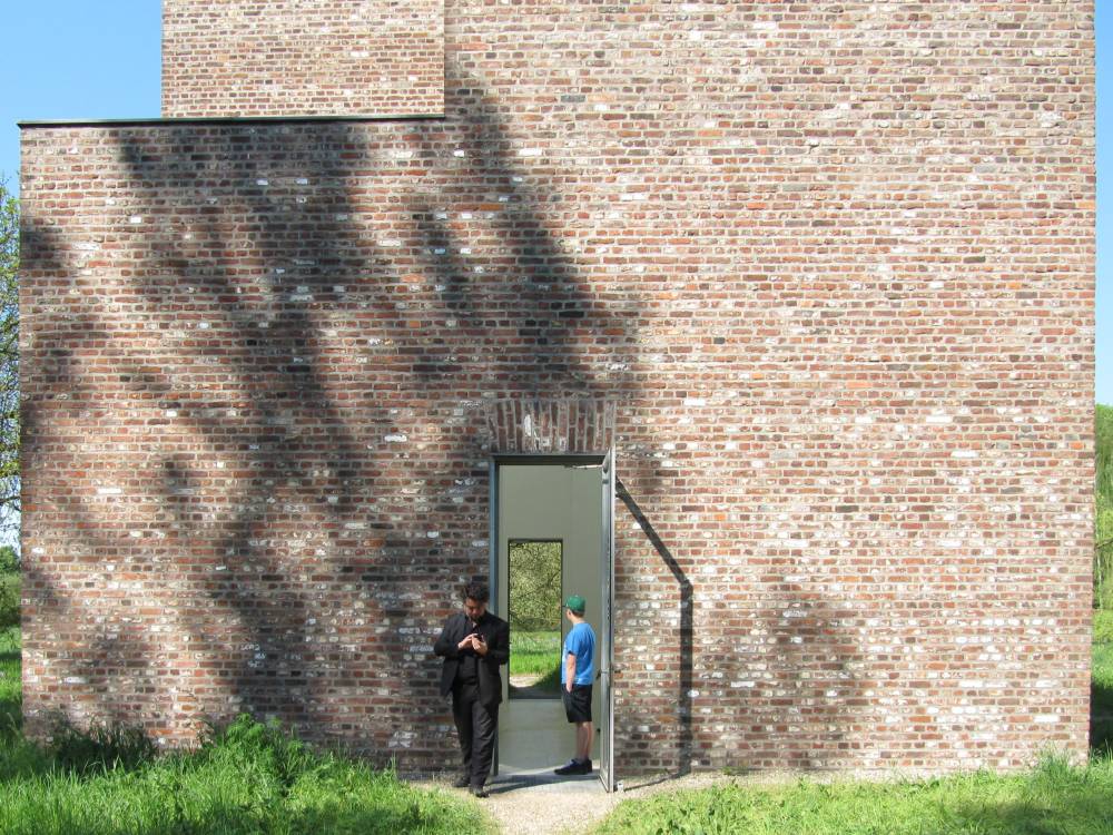 ich und das kind vor einem der bauten auf der museumsinsel hombroich. die türen des ziegelbaus sind offen, man kann durc beide türen hinter das gebäude schauen.