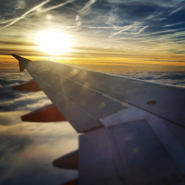An airplane wing above the clouds with the sun low on the horizon.
